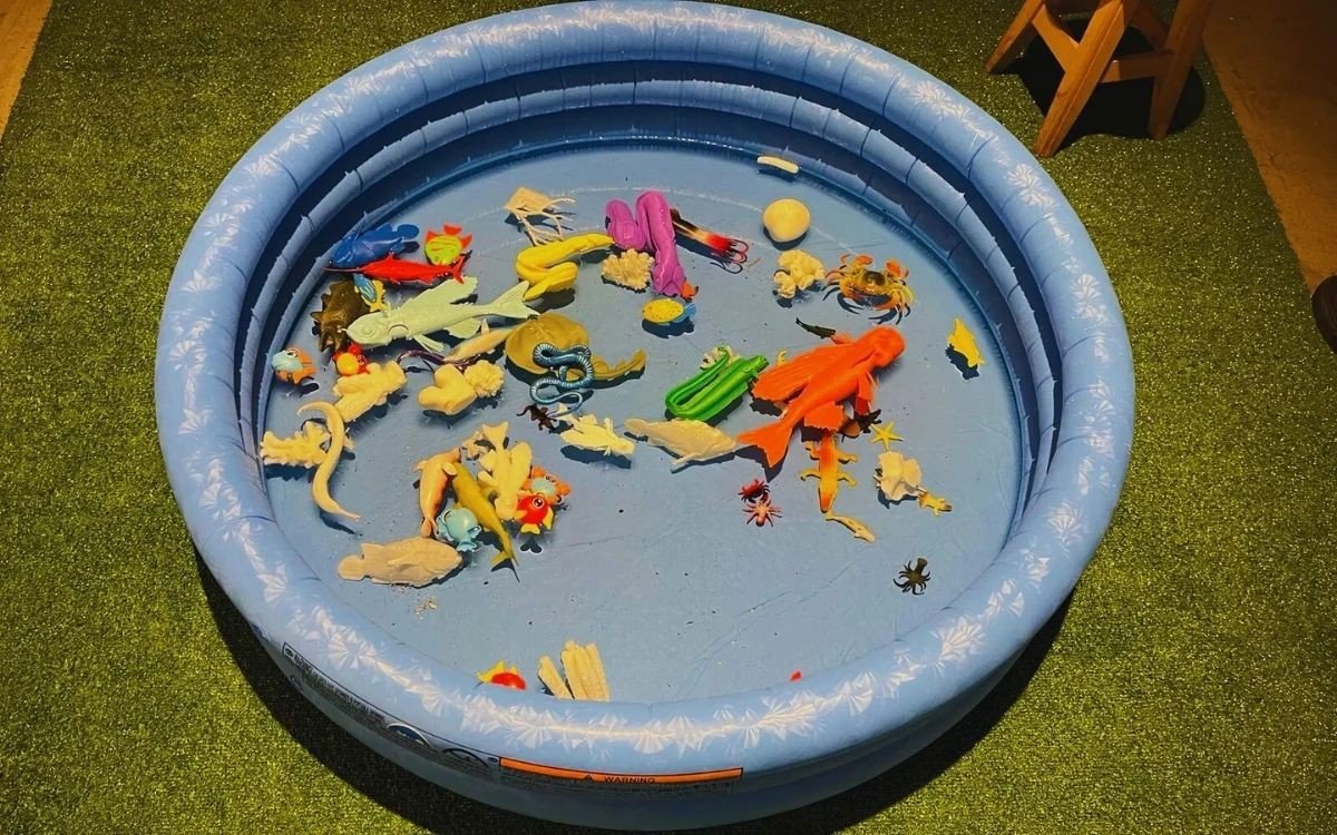 Детский надувной бассейн с разноцветными пластиковыми фигурками рыб и морских обитателей на зелёном полу галереи