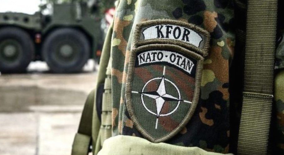 Нашивка KFOR NATO-OTAN на военной форме, на заднем плане бронетехника. Фото: Unknown