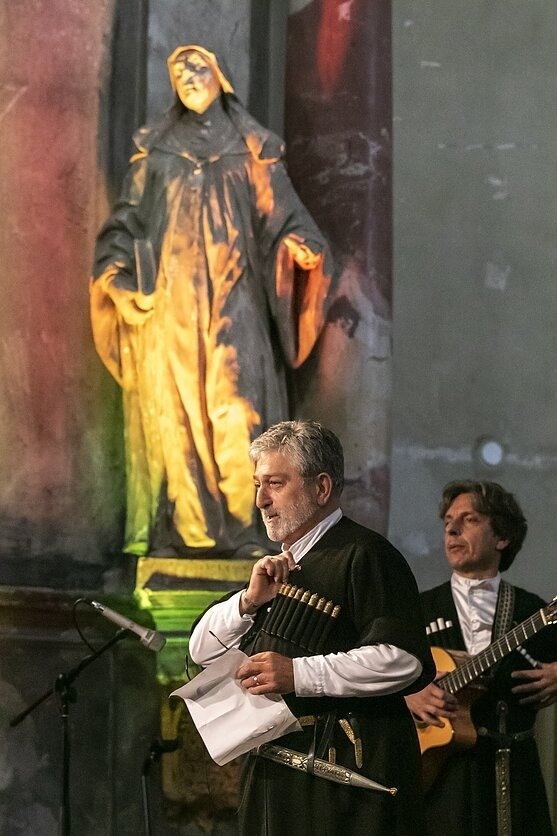 Солист в чохе читает текст, за ним гитарист, позади скульптура