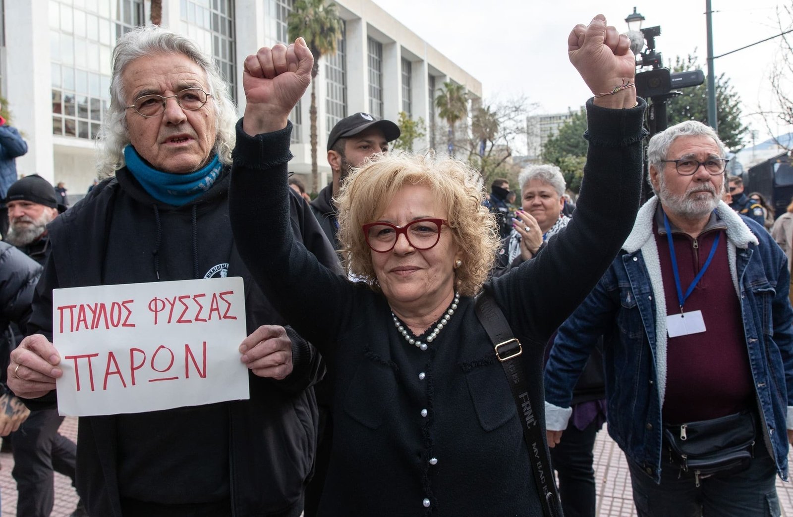Магда Фиссас, мать убитого неонацистами рэпера Павлоса Фиссаса, с поднятыми кулаками и плакатом «Павлос Фиссас — здесь» перед зданием суда
