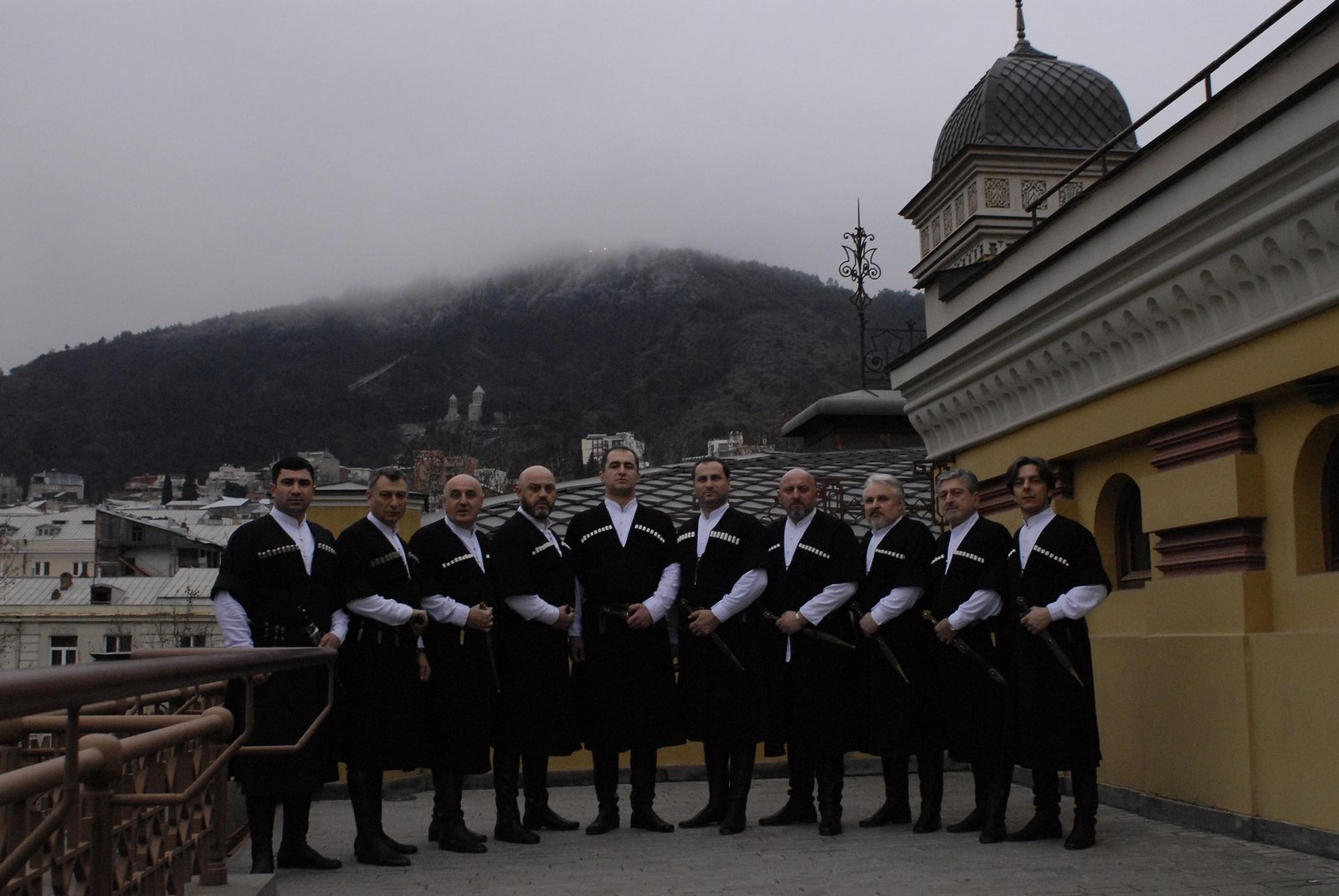 Группа мужчин в национальных чохах позирует на крыше с видом на Тбилиси