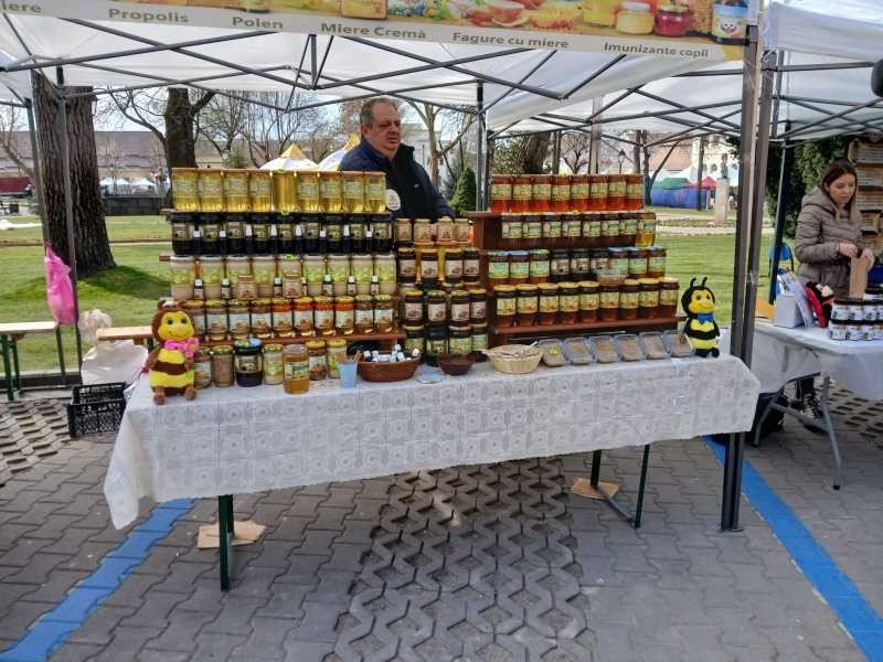 Мужчина за прилавком под навесом, перед ним ряды банок с мёдом, прополисом и пыльцой, рядом декоративные игрушки-пчёлки