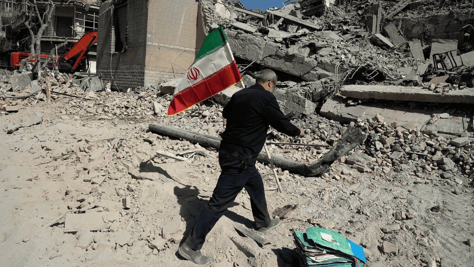 Мужчина с иранским флагом идёт среди руин разрушенного здания — последствия военных действий в Иране