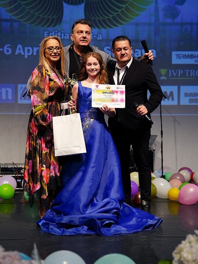 Церемония награждения на фестивале «Voci de Îngeri»: девочка в синем бальном платье с дипломом в окружении организаторов на сцене