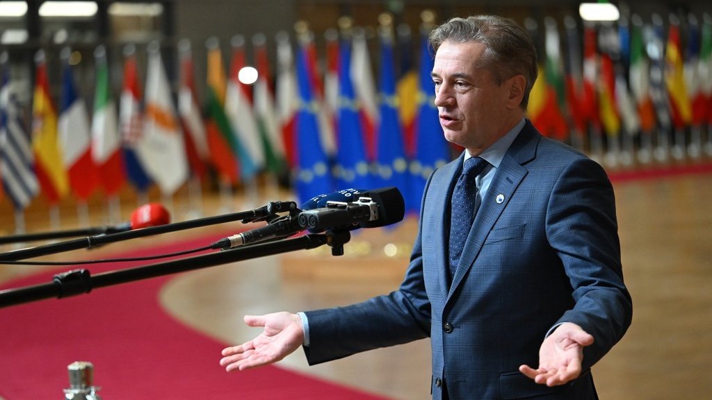 Роберт Голоб выступает перед журналистами на полях саммита Европейского совета, жестикулируя на фоне флагов стран-членов ЕС