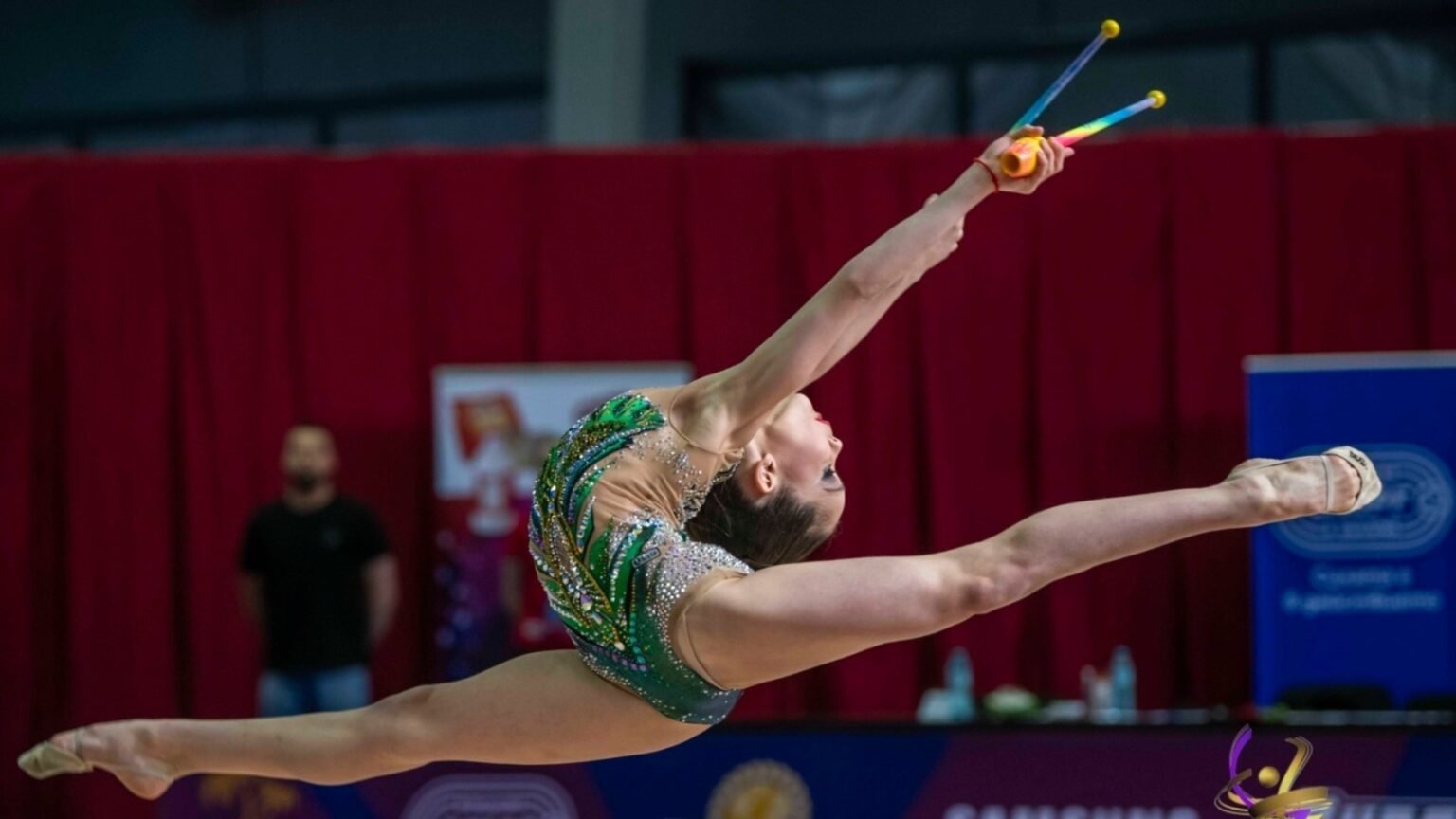 Болгарская гимнастка выполняет эффектный прыжок в шпагат с булавами на международном турнире по художественной гимнастике