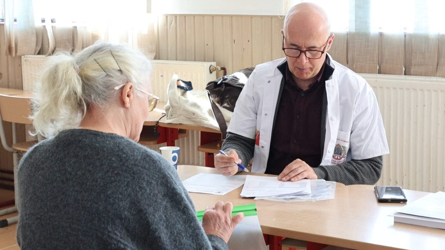 Врач в белом халате проводит консультацию пожилой женщины за столом в рамках выездной медицинской кампании
