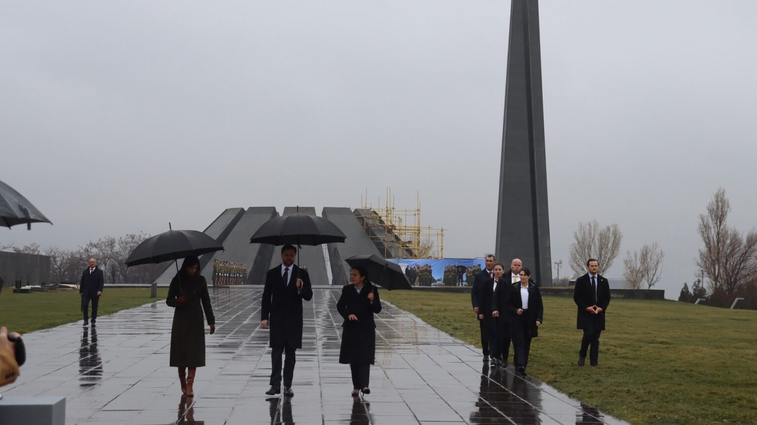 Делегация с зонтами идёт по дождливой аллее мемориального комплекса Цицернакаберд в Ереване