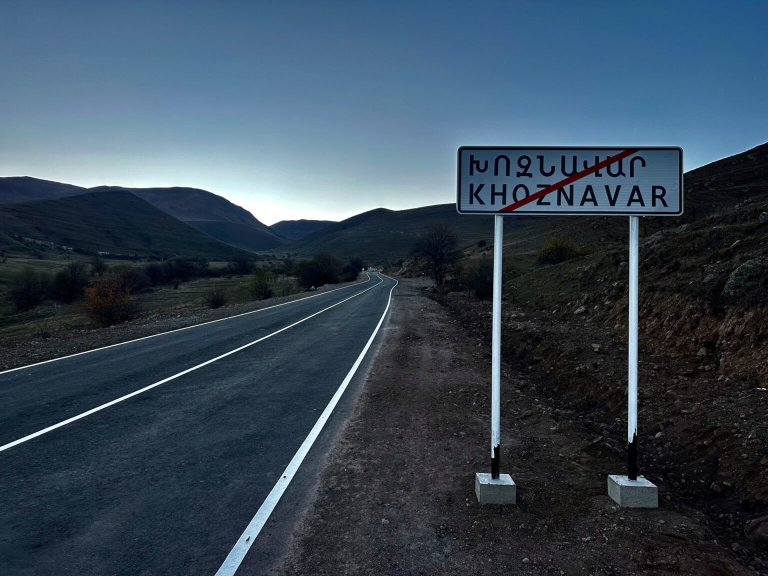 Дорожный знак на выезде из села Хознавар в Сюникской области Армении — пустое шоссе уходит вдаль среди гор на закате