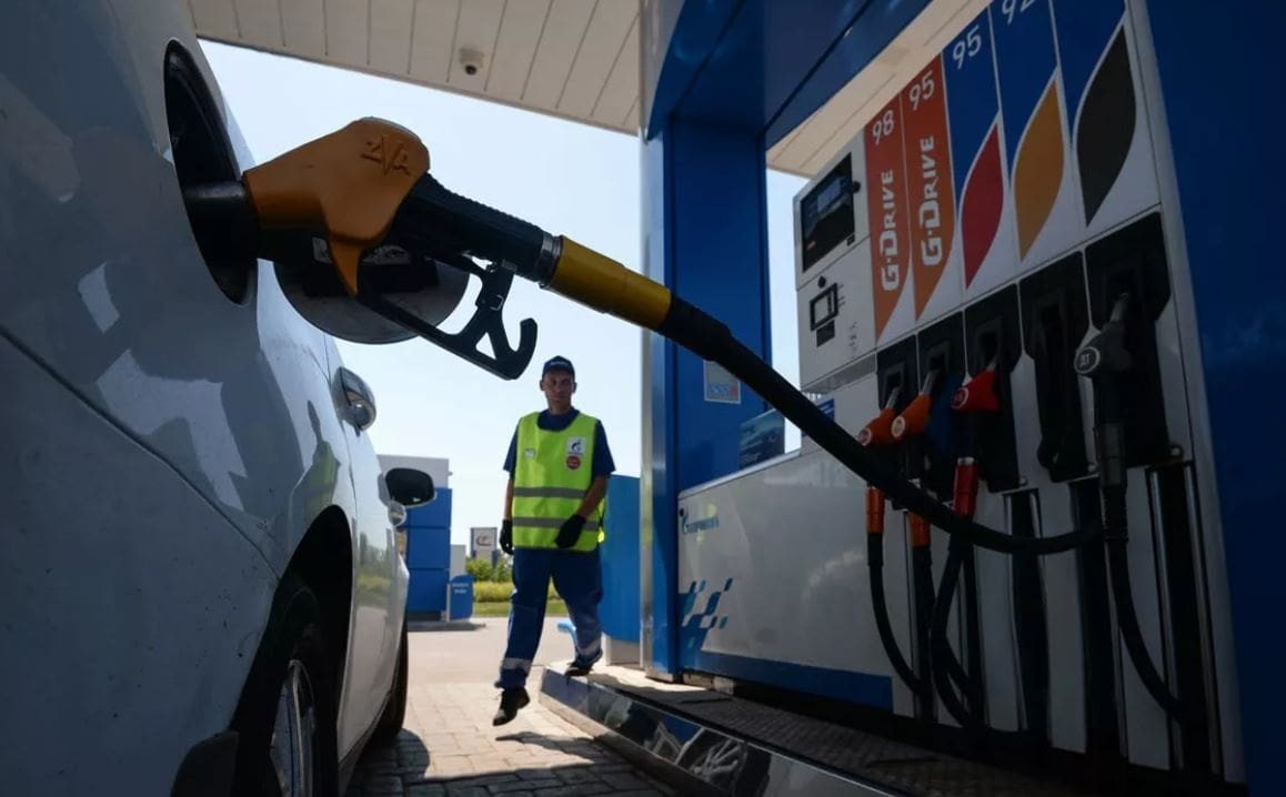 Заправка автомобиля на АЗС — топливный пистолет вставлен в бак, на заднем плане сотрудник станции в светоотражающем жилете