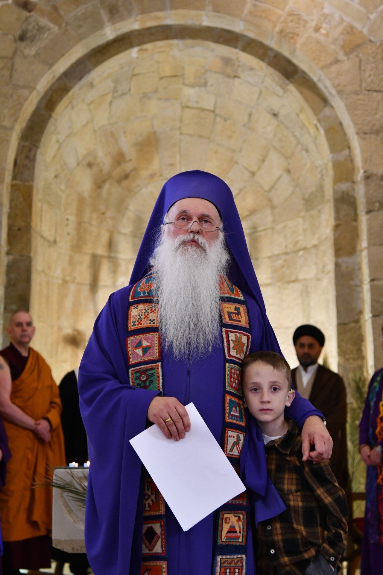 Епископ Малхаз Сонгулашвили в фиолетовом облачении стоит с ребёнком в арочном проёме Кафедрального собора мира, на заднем плане видны представители буддийского и мусульманского духовенства