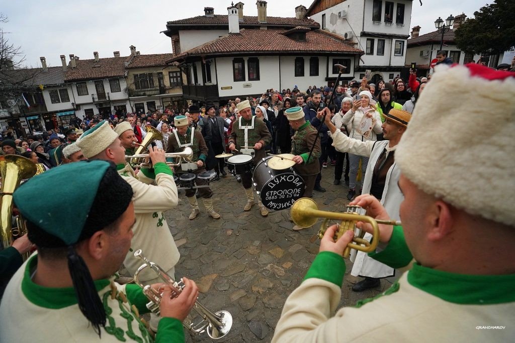 Духовой оркестр в зелёных мундирах играет на площади перед толпой горожан на фоне традиционных тырновских домов