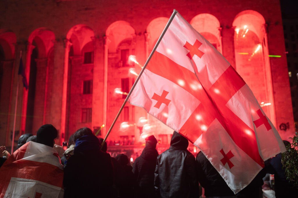 Протестующие грузинской оппозиции с национальными флагами во время ночной демонстрации в Тбилиси на фоне правительственного здания, освещённого красными сигнальными огнями
