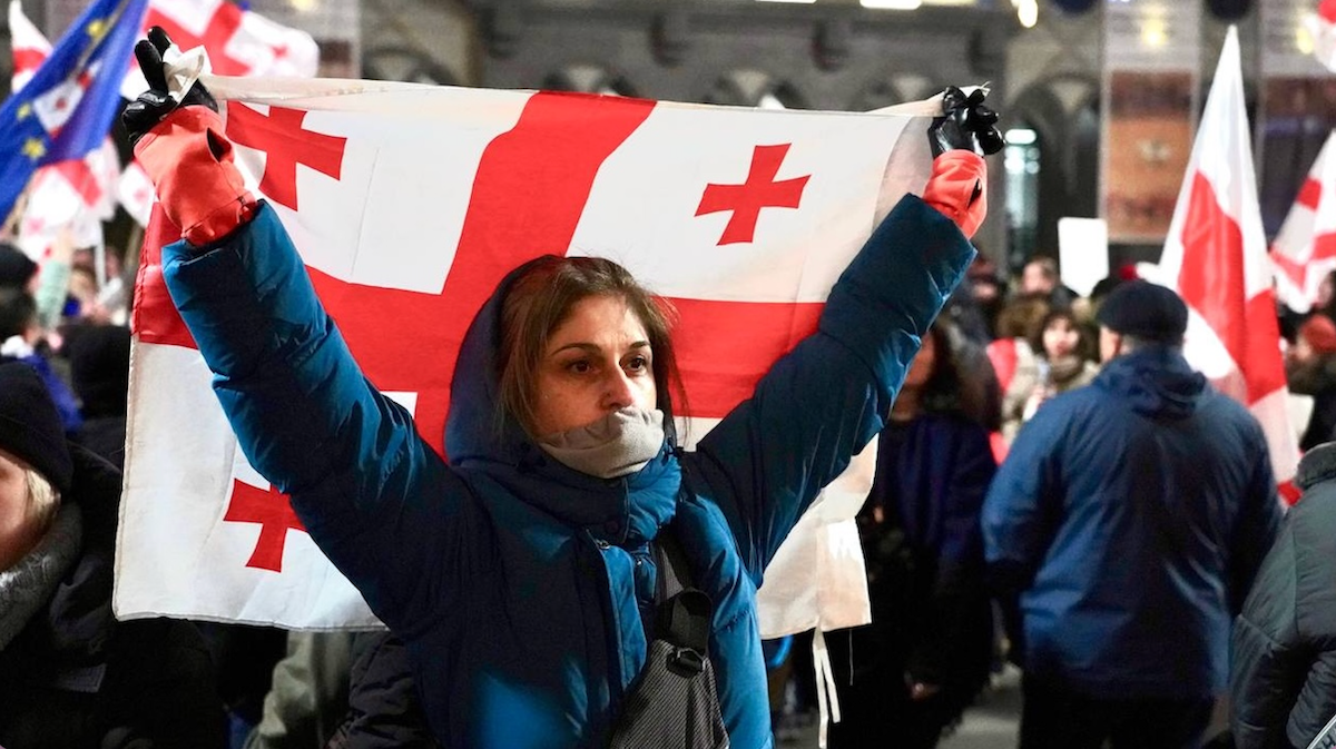 Молодая женщина в синей куртке держит грузинский флаг над головой во время протеста, на заднем плане виден флаг ЕС, за ней толпа протестующих. Фото: JAM News