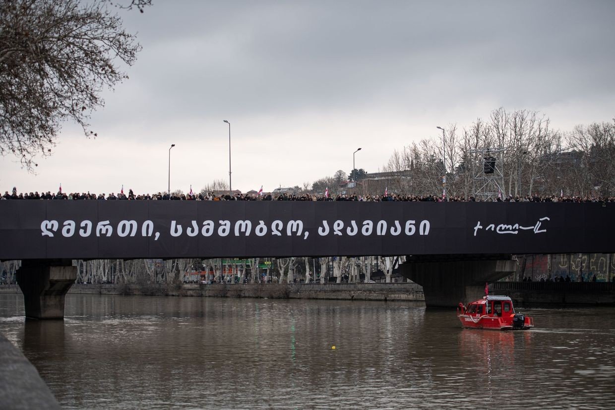 Мемориальный баннер с надписью на грузинском языке и подписью Патриарха Илии II на мосту через реку Кура в Тбилиси; на мосту — тысячи людей с флагами