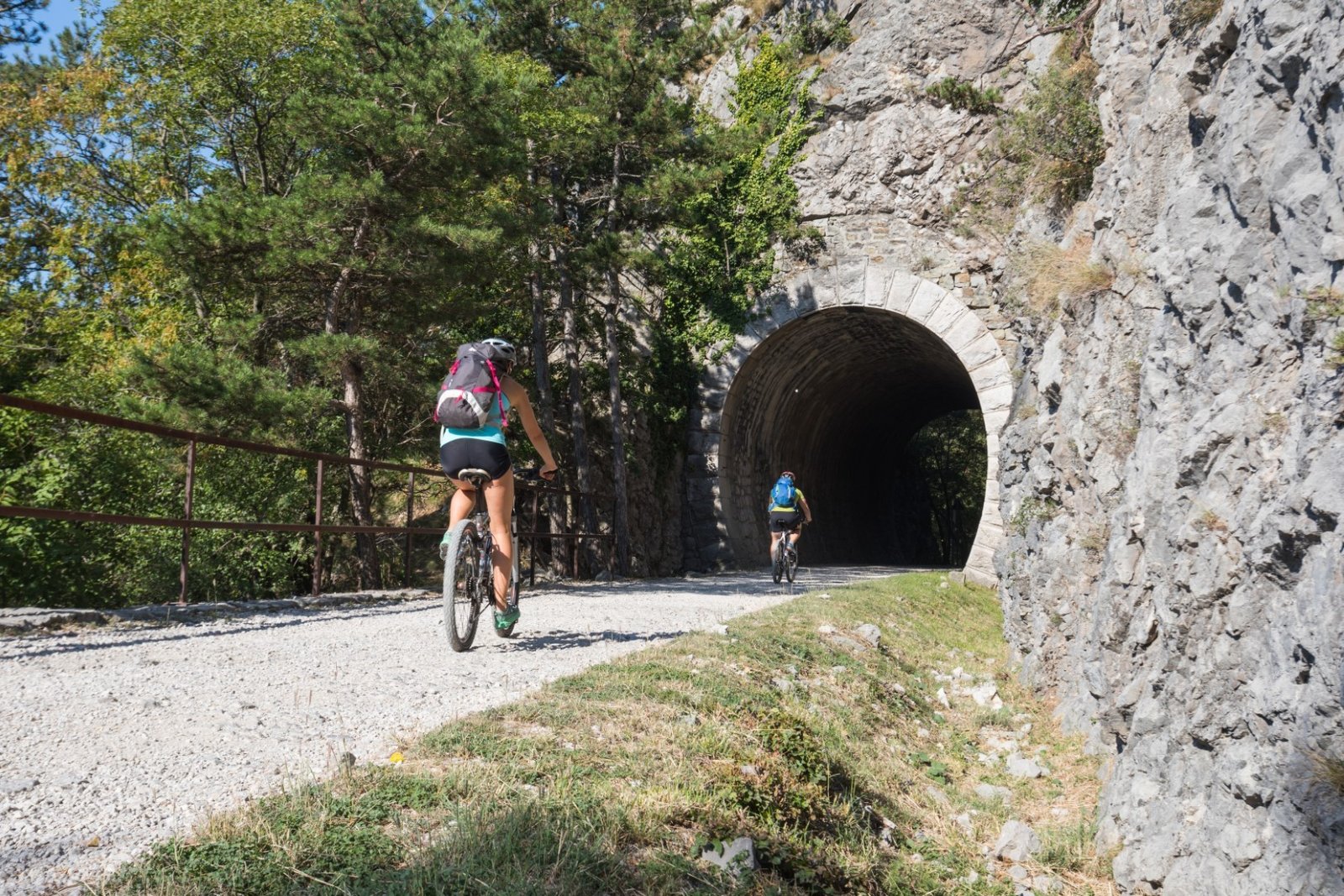 Велосипедисты въезжают в скальный тоннель на гравийной тропе в долине Глинщица, окружённой утёсами и соснами