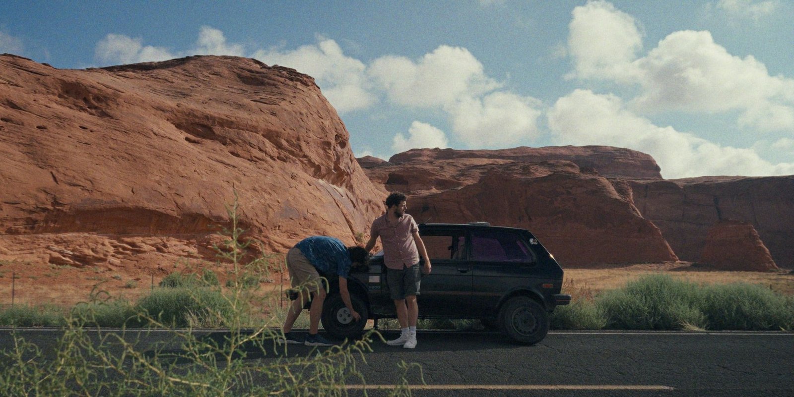 Участники путешествия ремонтируют Yugo на обочине дороги на фоне красных скал в пустыне на юго-западе США — кадр из фильма «Yugo Goes to America»
