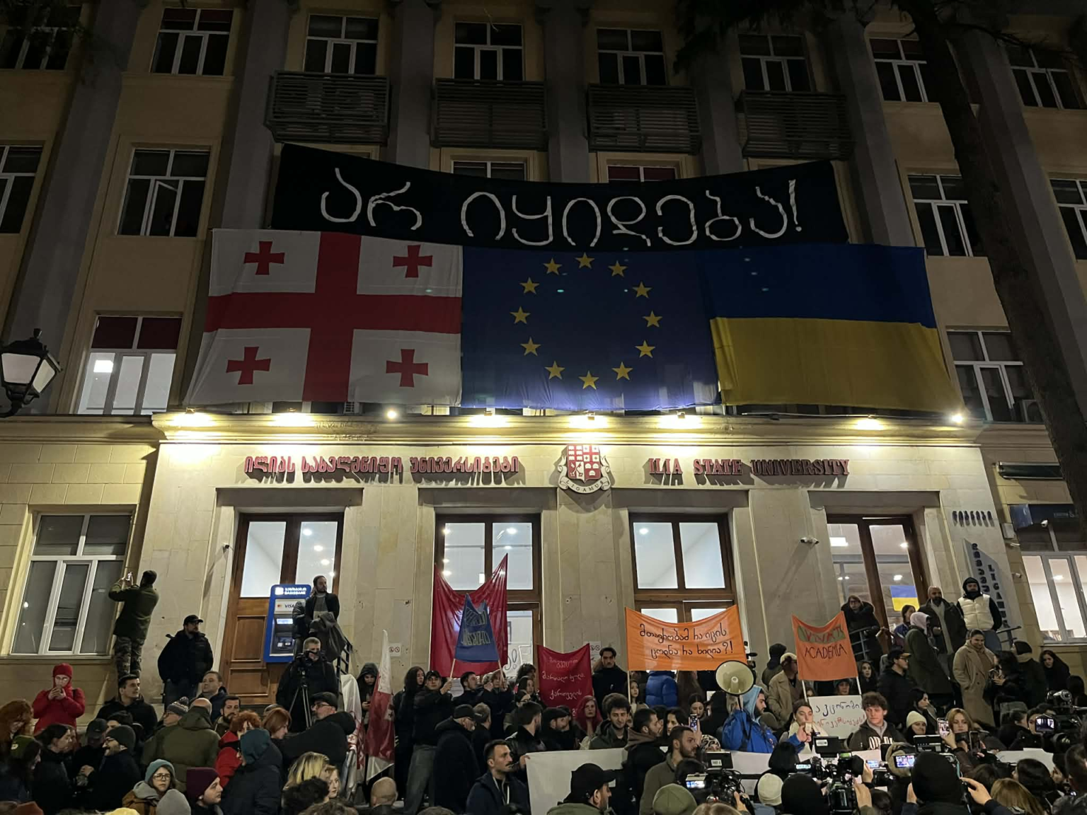 Ночной протест у здания Государственного университета Ильи в Тбилиси — на фасаде вывешены флаги Грузии, Евросоюза и Украины, внизу собрались участники акции с транспарантами