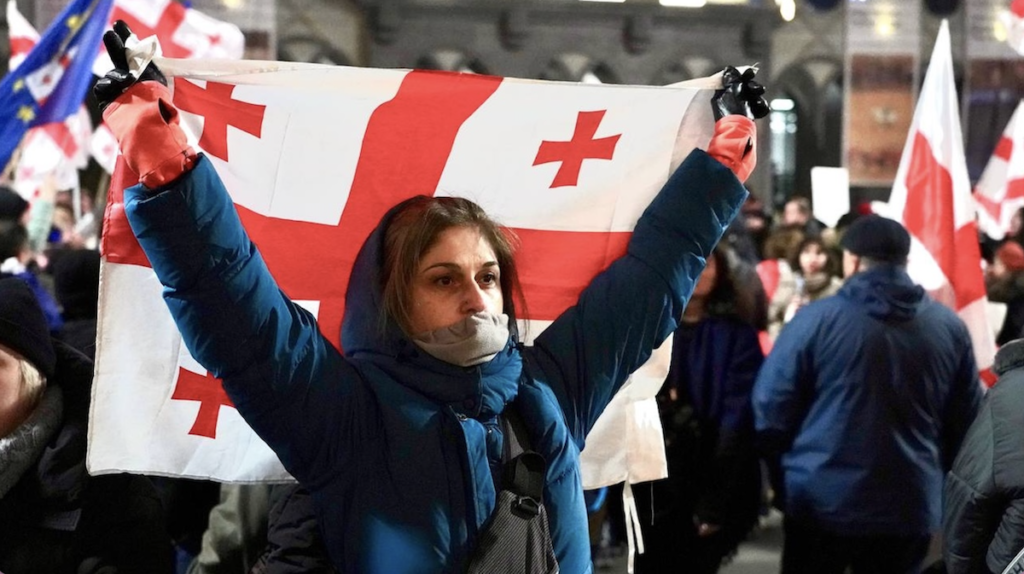 Участник протестной акции держит грузинский национальный флаг на ночном митинге против репрессивного законодательства, на заднем плане видны флаги ЕС