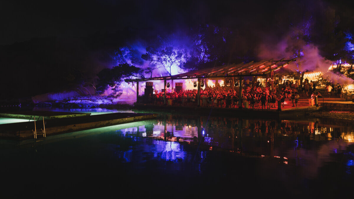 Ночной вид на площадку The Garden Resort в Тисно — фиолетовые и синие прожекторы отражаются в воде, создавая атмосферный пейзаж