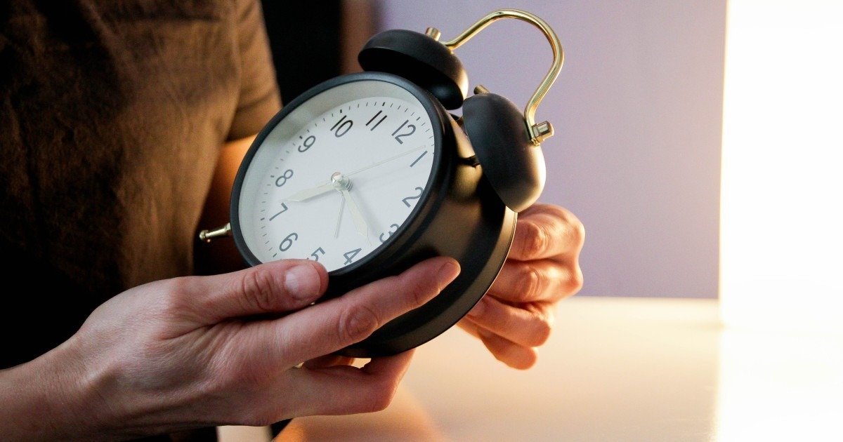 Человек держит в руках чёрный будильник в приглушённом вечернем свете