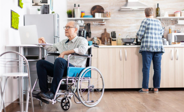 Пожилой мужчина в инвалидном кресле работает за ноутбуком на кухне, пока женщина готовит рядом