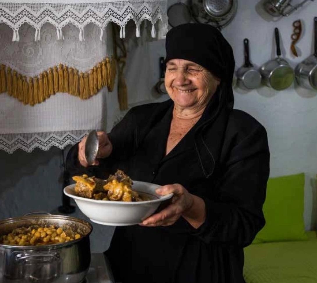 Критская женщина в традиционном чёрном одеянии готовит домашнее блюдо на деревенской кухне