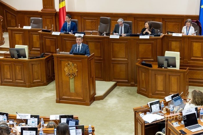 Заседание парламента Молдовы: спикер выступает с трибуны на фоне флагов ЕС и Молдовы, в зале — делегаты и чиновники