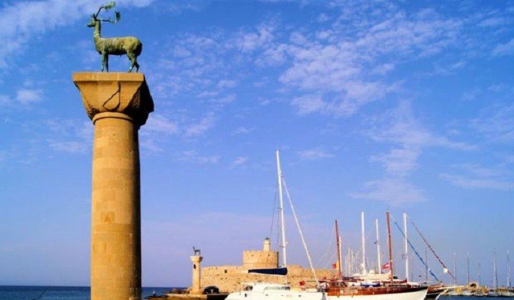Колонна с бронзовым оленем у входа в порт Мандраки на Родосе, на заднем плане — яхты и крепость Святого Николая