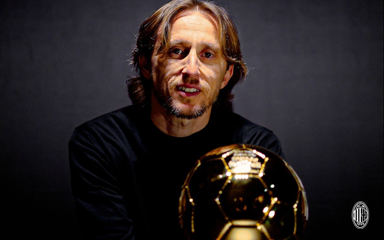 Крупный портрет Луки Модрича за «Золотым мячом», футболист смотрит в камеру, в нижнем правом углу видна эмблема «Милана»