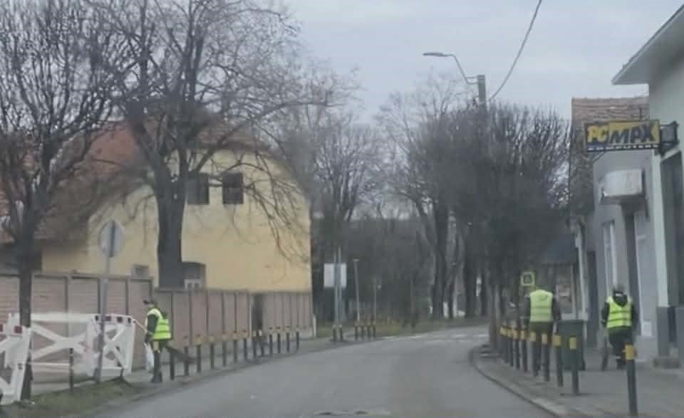 Улица в Сремска-Митровице в пасмурную погоду — голые деревья и рабочие в жёлтых жилетах на тротуаре