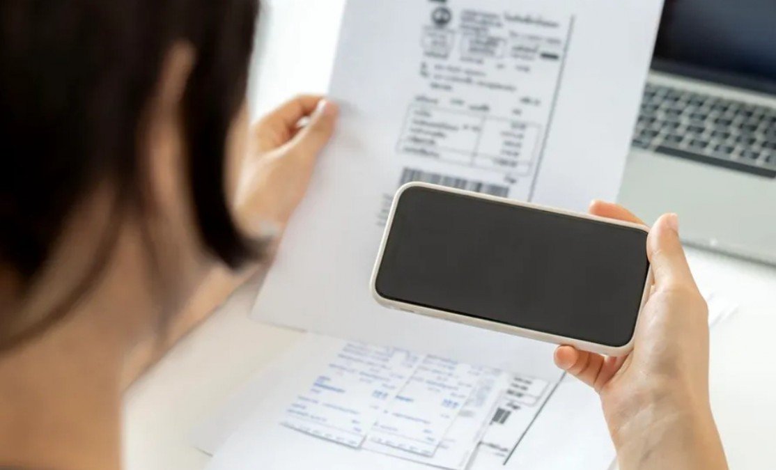Женщина изучает счёт за коммунальные услуги, держа в руках смартфон перед ноутбуком