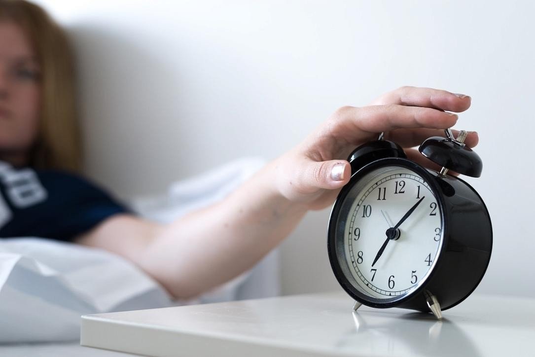 Женщина в постели тянется рукой к звонящему будильнику на прикроватной тумбочке