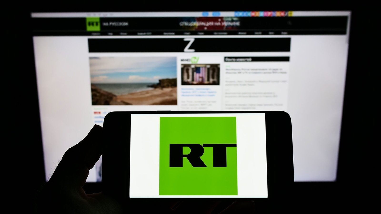 Логотип RT (Russia Today) на экране смартфона на фоне сайта RT на мониторе — иллюстрация присутствия российских государственных СМИ