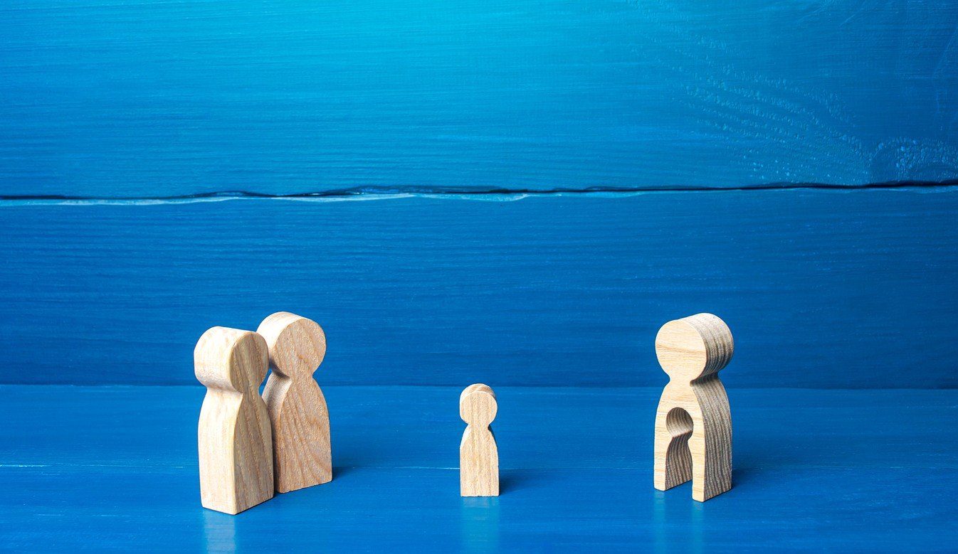 Деревянные фигурки на синем фоне, изображающие пару, ребёнка и беременную женщину с пустотой внутри, символизирующие суррогатное материнство