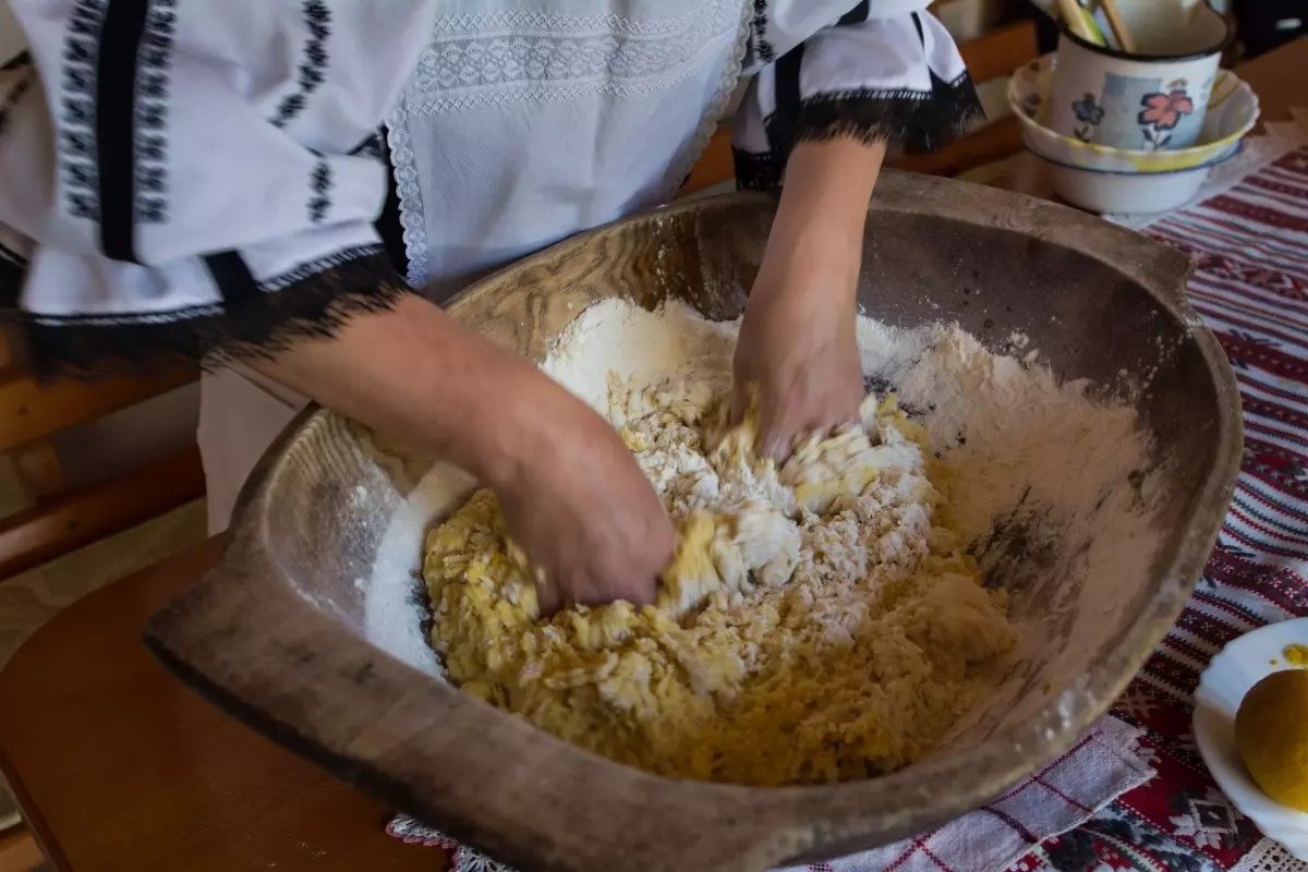 Женщина в традиционной румынской вышиванке замешивает тесто руками в большой деревянной миске