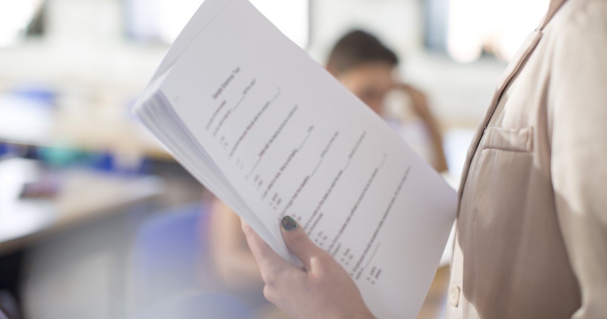 Преподаватель держит в руках экзаменационные бланки на фоне учебной аудитории