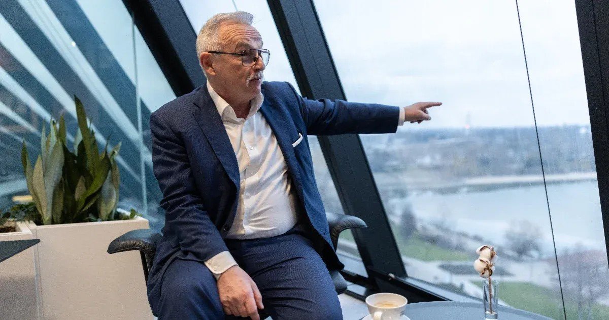 Business executive in navy suit and glasses sitting by large office windows, pointing outward. Фото: Telex