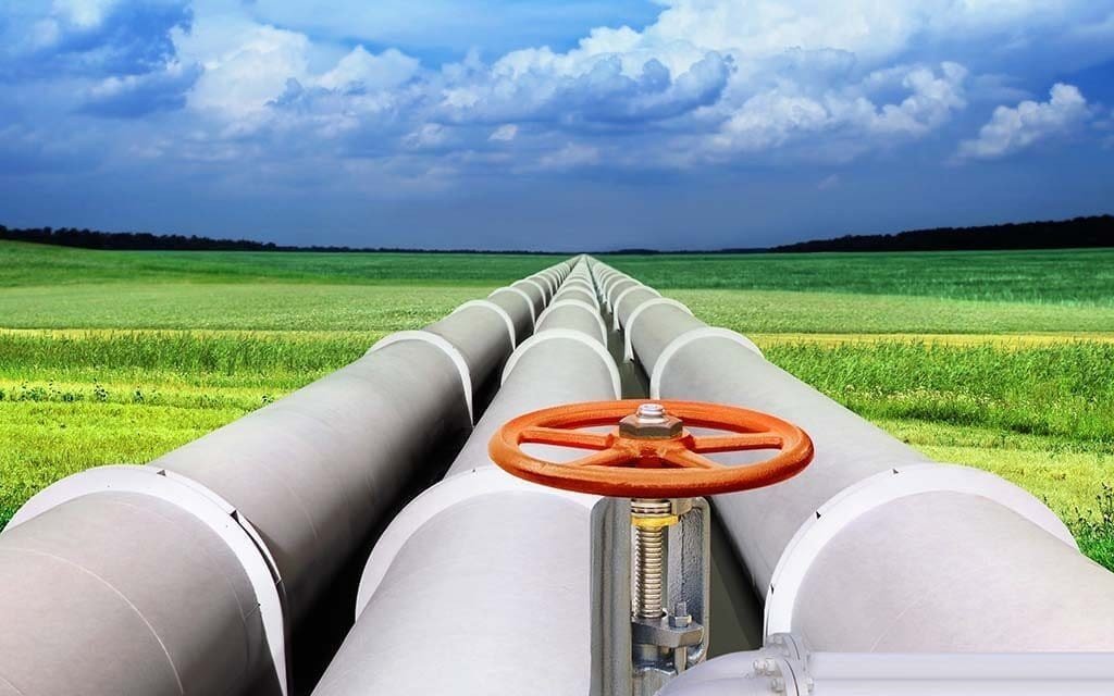Oil pipeline with orange valve extending through green fields under dramatic cloudy sky. Фото: Tozsdeforum