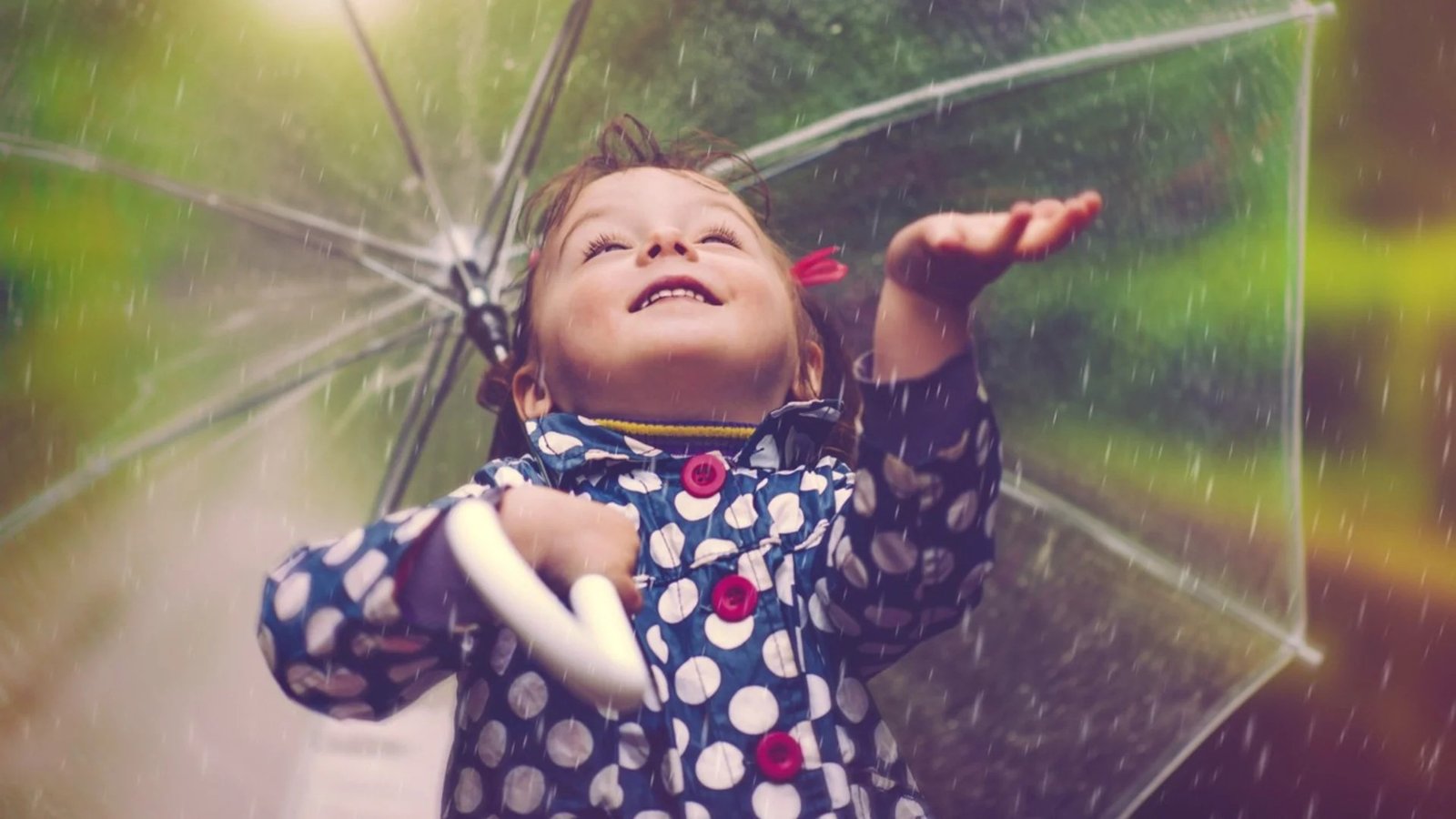 Девочка в плаще в горошек радуется дождю, протягивая руку из-под прозрачного зонта
