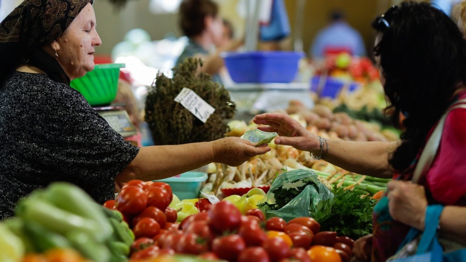 Женщина-продавщица передаёт сдачу покупательнице за прилавком с помидорами, перцем и зеленью на открытом рынке в Румынии