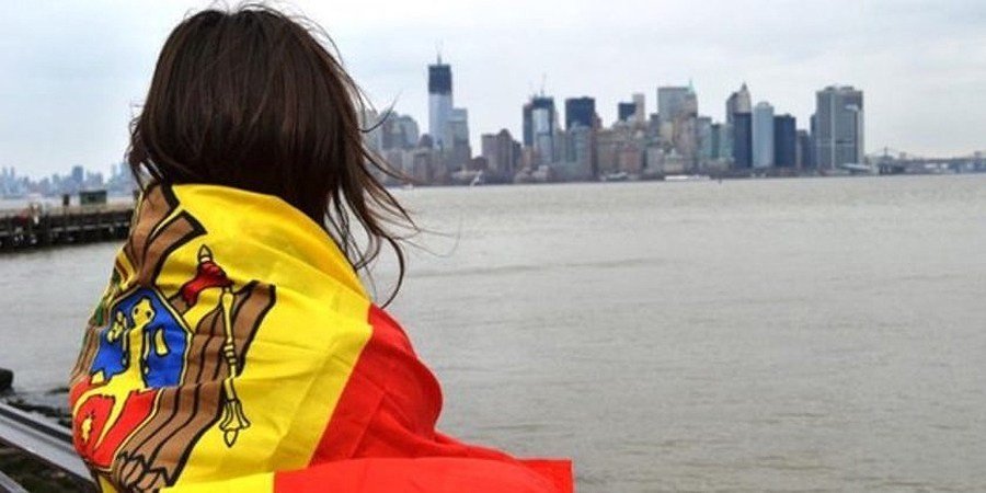 Молодая женщина, закутанная в молдавский триколор, смотрит на небоскрёбы Манхэттена за рекой — визуальный символ массового оттока молодёжи и рабочей силы из Молдовы за рубеж