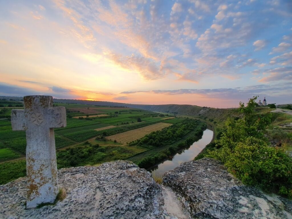 Каменный средневековый крест на краю скального плато Старого Орхея; внизу — излучина реки Реут, зелёные сельские угодья и церковь на закате
