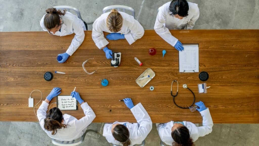 Вид сверху на медицинских работников в белых халатах и перчатках за рабочим столом: микроскоп, стетоскоп, пробирки и записи по результатам анализов