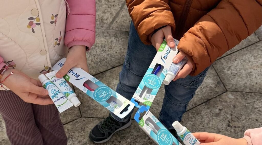 Двое детей в зимних куртках держат в руках упаковки зубных щёток и тюбики паст Sensodyne и Parodontax, полученные на площадке Каравана здоровья полости рта в одном из городов Румынии