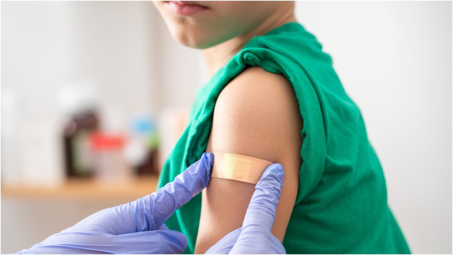 Ребёнок в зелёной футболке с пластырем на плече после прививки ROR — медработник в синих перчатках фиксирует пластырь