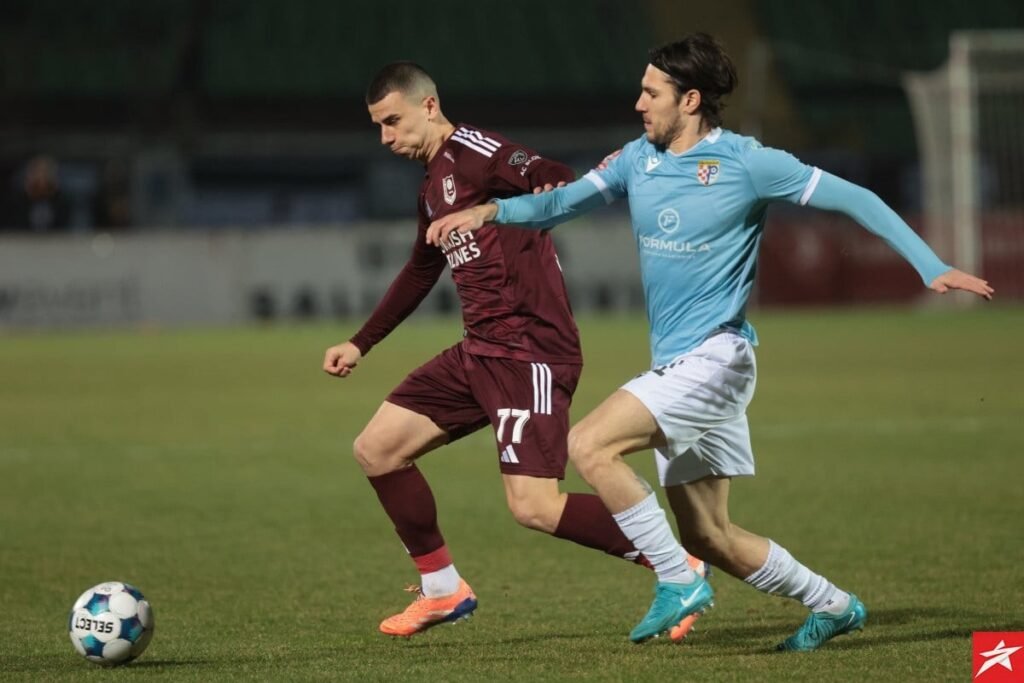 Игрок ФК «Сараево» в бордовой форме с номером 77 ведёт мяч под прессингом соперника в светло-голубой форме на травяном поле боснийской WWin лиги