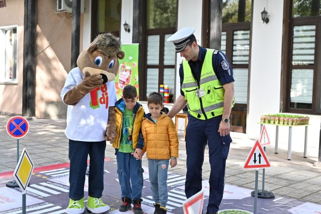 Талисман кампании TEDI и сотрудник дорожной полиции переводят двух дошкольников через размеченный пешеходный переход во дворе детского сада в Пловдиве