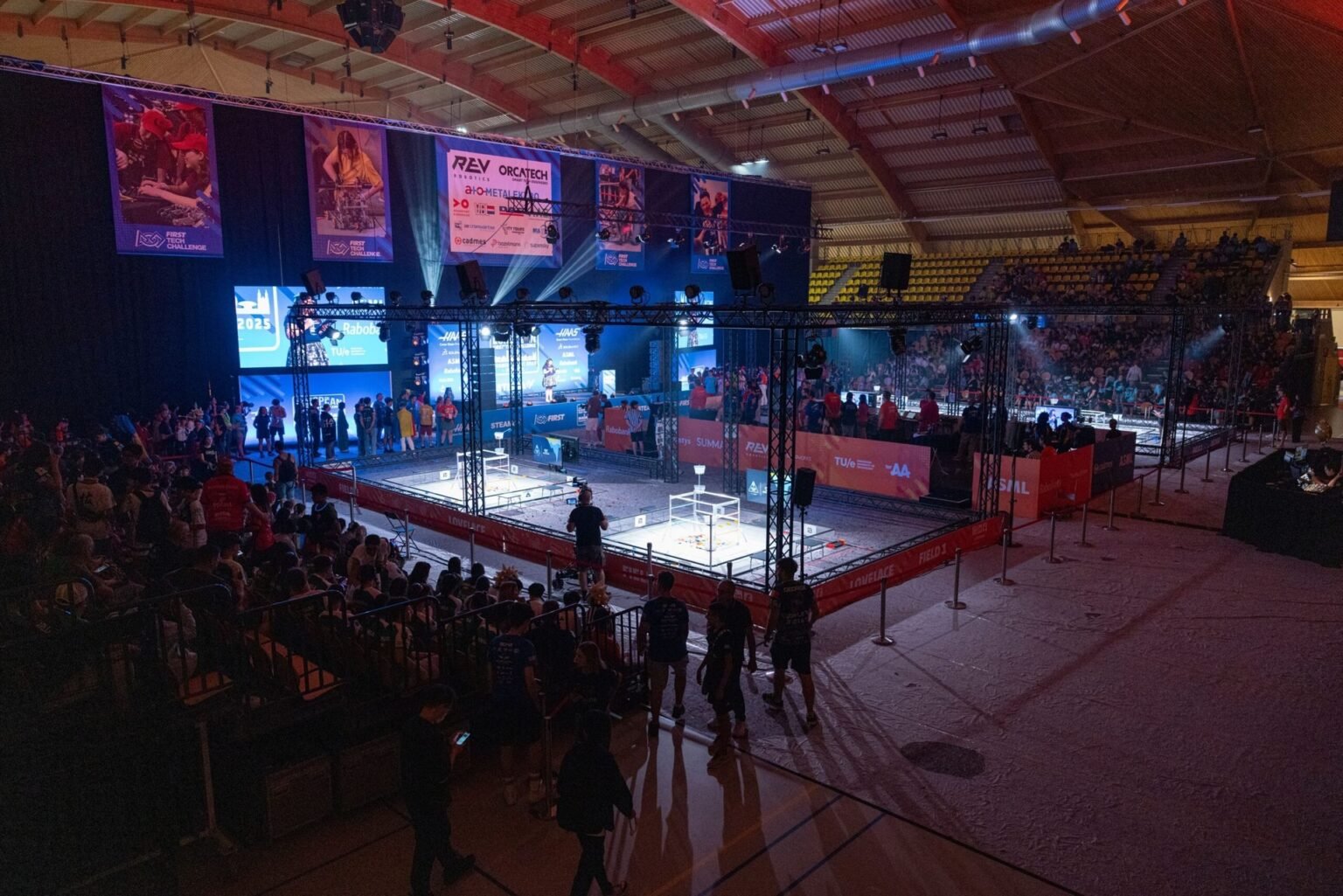 Общий вид арены FIRST Tech Challenge: две огороженные площадки с роботами-участниками в центре зала, яркая подсветка, большие экраны с логотипами FIRST, REV и Orcatech, заполненные зрителями трибуны