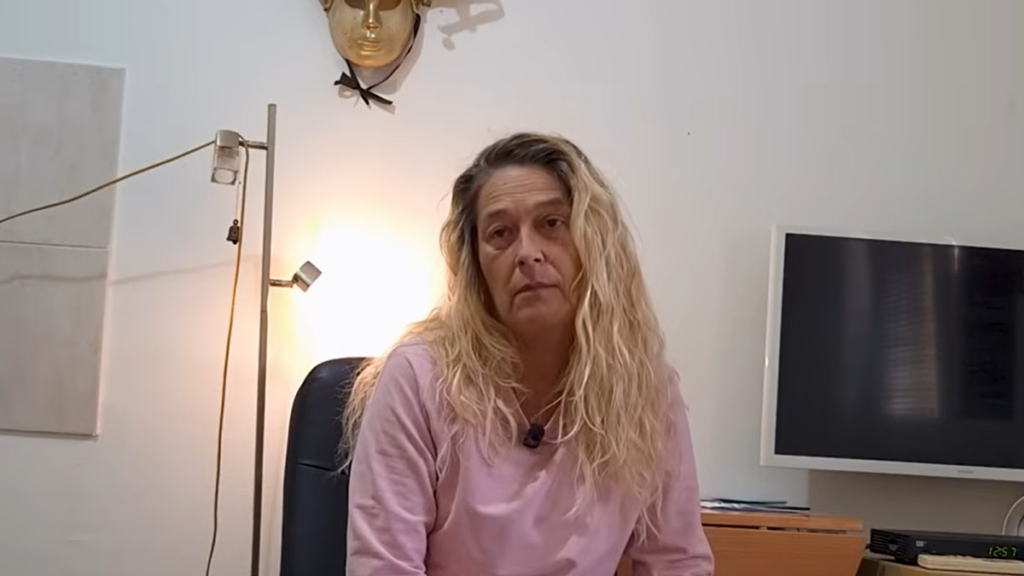 Женщина со светлыми волнистыми волосами в розовой кофте сидит в небольшой комнате на фоне белой стены с венецианской маской и напольной лампой